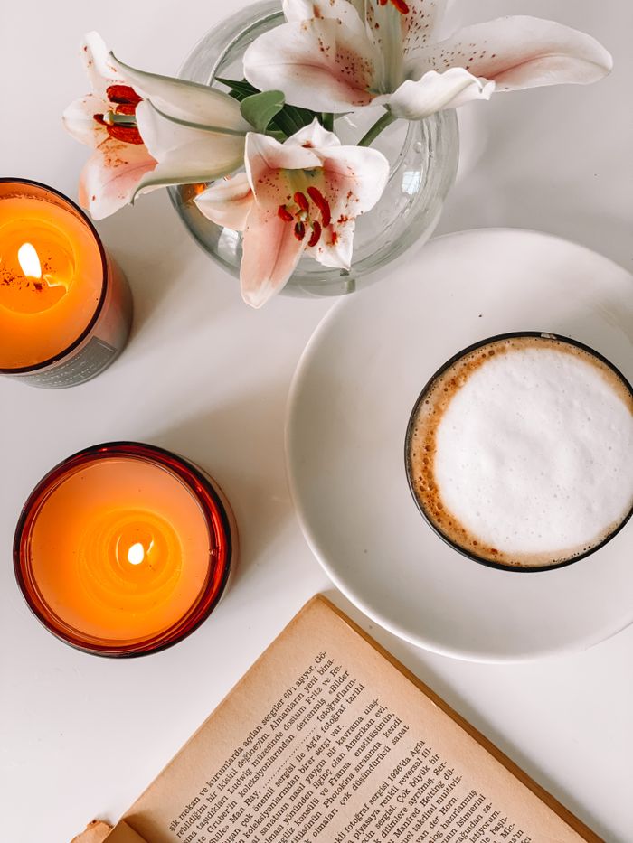 A warm ambiance with candles, a cup of coffee, flowers, and a book on a white surface.